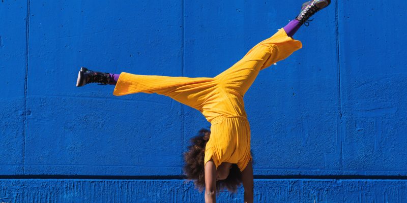 Full body of African American teen girl in bright yellow jumpsuit performing handstand against blue wall on street
